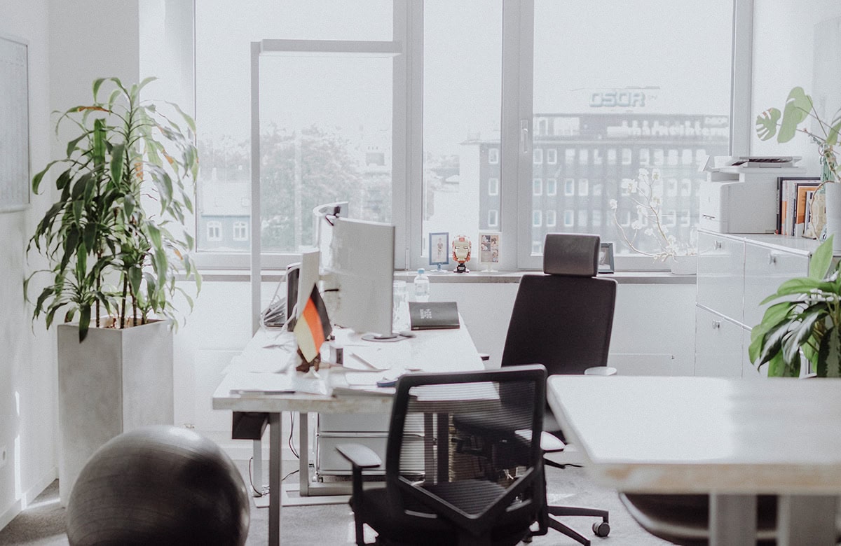 Modernes Bürozimmer mit Pflanzen, grauen Möbeln, und vielen Fenstern. Wenige persönliche Gegenstände.