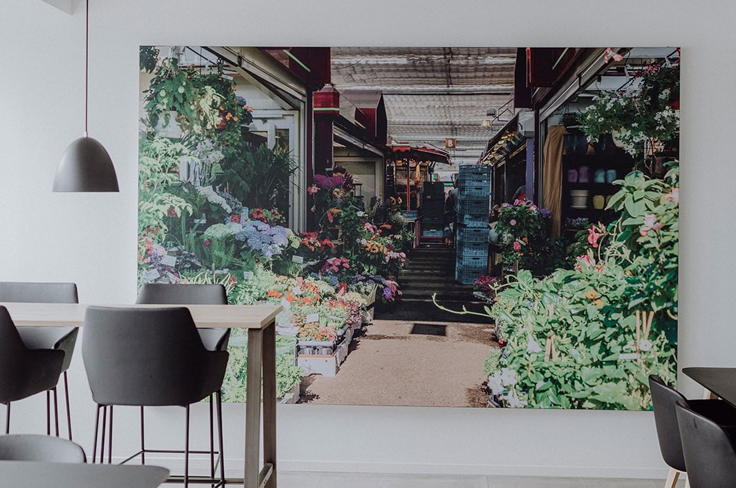 Ein großes Gemälde eines lebhaften Blumenmarkts, umgeben von schwarzen Stühlen und weißen Wänden.